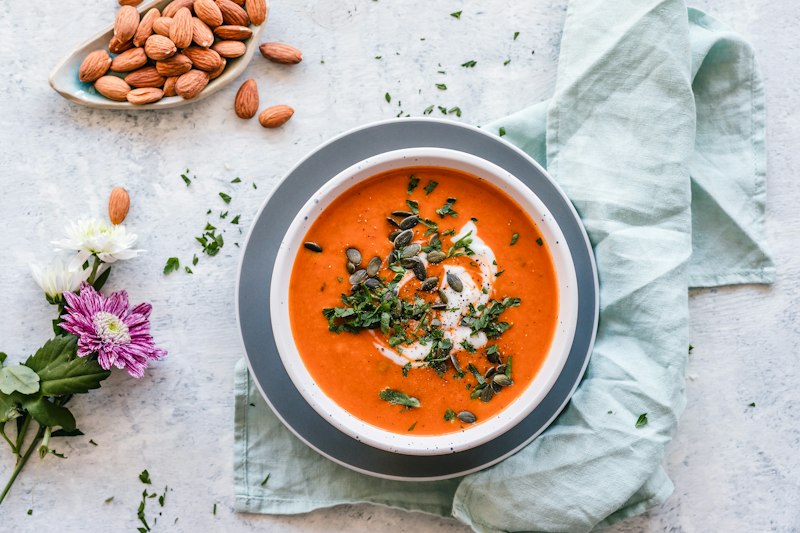 Orange soup topped with seeds and herbs on pale green linen beside flowers and almonds